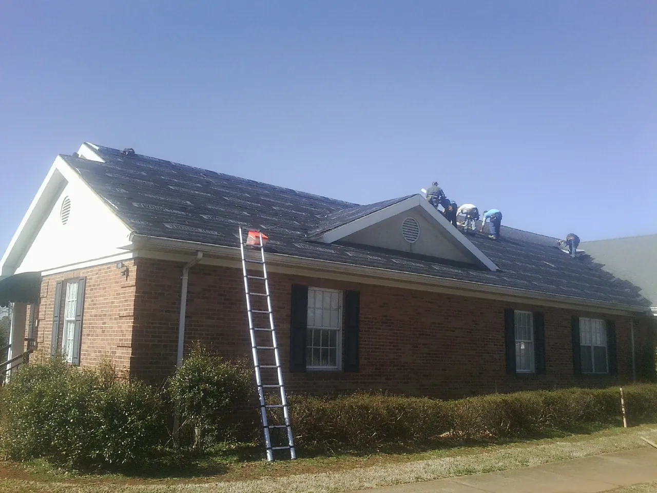 Crew tearing shingles off a brick ranch roof with a ladder set — Emergency Roof Repair work by our crew in Claridge