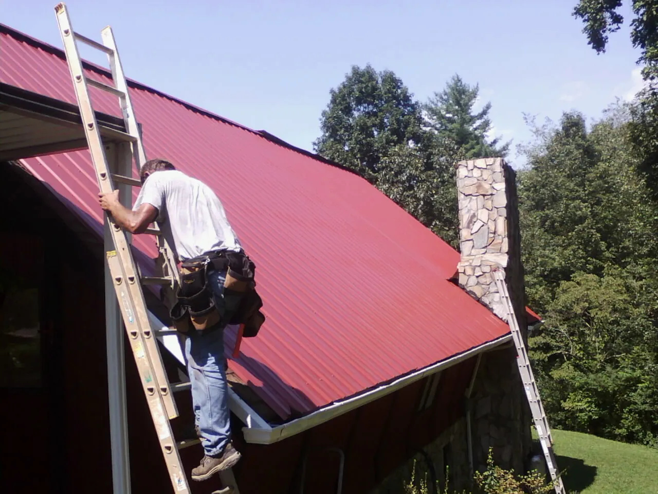 Roofer on a ladder beside a red metal roof and stone chimney — Soffit & Fascia Repair by Roofing Brothers in Claridge